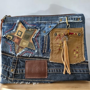 May include: A denim clutch with a patchwork star design and a brown leather tassel. The clutch features a brown leather patch with the Hollister logo. The bag has a zipper closure and a brown leather strap.