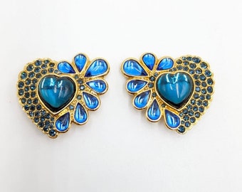 May include: A pair of gold-tone heart-shaped earrings with blue crystal accents. The earrings feature a large blue crystal in the centre of each heart.