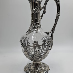 May include: A silver and glass decanter with an ornate design. The decanter has a clear glass body and a silver base and handle. The handle is shaped like a vine with leaves and flowers.