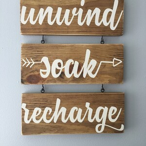 May include: Three wooden signs with white lettering hanging from a wall. The top sign says "unwind", the middle sign says "soak" with an arrow on either side, and the bottom sign says "recharge".