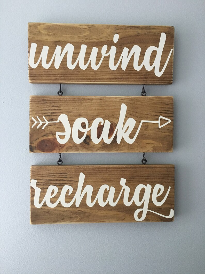 May include: Three wooden signs with white lettering hanging from a wall. The top sign says "unwind", the middle sign says "soak" with an arrow on either side, and the bottom sign says "recharge".