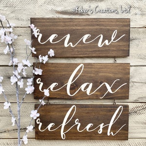 May include: Three wooden signs with white lettering hanging on a white wood wall. The signs read "renew", "relax", and "refresh".