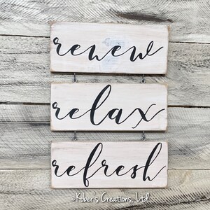 May include: Three white wooden signs with black cursive lettering hanging on a wood wall. The signs read "renew", "relax", and "refresh".