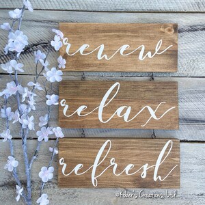 May include: Three wooden signs with white lettering hanging on a white wood wall. The signs read "renew", "relax", and "refresh".