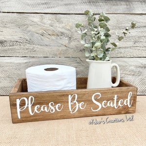 May include: A wooden box with the text "Please Be Seated" in white lettering. The box is stained a dark brown and has a roll of toilet paper and a white pitcher with greenery inside.