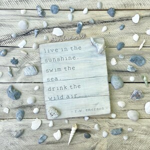 May include: A white wooden sign with a quote by R.W. Emerson: "live in the sunshine, swim the sea, drink the wild air." The sign is surrounded by seashells and stones on a wooden surface.