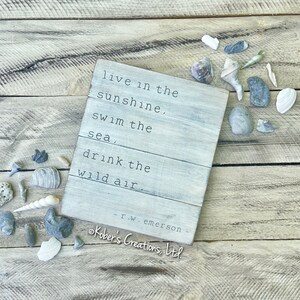 May include: A white wooden sign with a quote by R.W. Emerson that reads "live in the sunshine, swim the sea, drink the wild air". The sign is on a wooden background with seashells scattered around it.