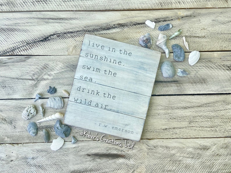 May include: A white wooden sign with a quote by R.W. Emerson that reads "live in the sunshine, swim the sea, drink the wild air". The sign is on a wooden background with seashells scattered around it.