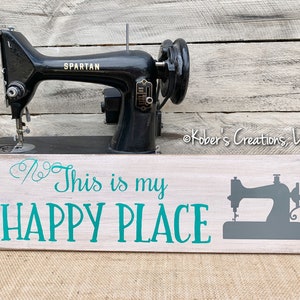 May include: A wooden sign with white paint and a teal green inscription that reads "This is my HAPPY PLACE". A gray silhouette of a sewing machine is on the right side of the sign.