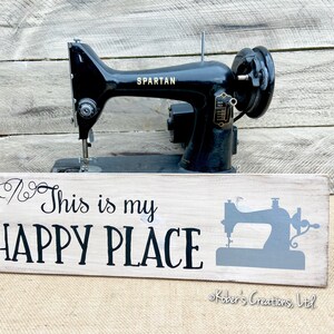 May include: A white wooden sign with black text that reads "This is my HAPPY PLACE" and a gray silhouette of a sewing machine. The sign is sitting in front of a vintage black sewing machine.