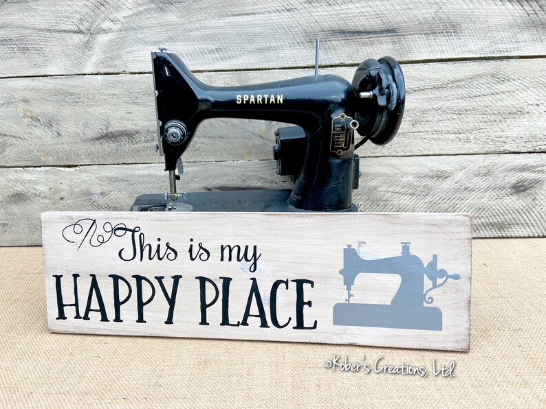 May include: A white wooden sign with black text that reads "This is my HAPPY PLACE" and a gray silhouette of a sewing machine. The sign is sitting in front of a vintage black sewing machine.
