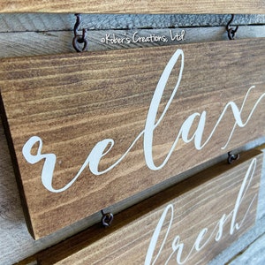 May include: Two wooden signs with white lettering hanging on a wall. The top sign says "relax" and the bottom sign says "refresh".