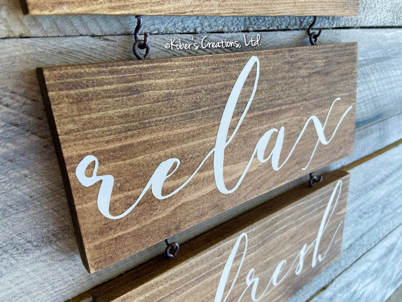 May include: Two wooden signs with white lettering hanging on a wall. The top sign says "relax" and the bottom sign says "refresh".