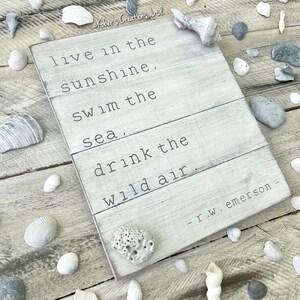 May include: White wooden sign with a quote about living in the sunshine, swimming in the sea, and drinking the wild air. The quote is attributed to R.W. Emerson.
