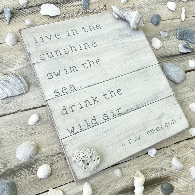 May include: White wooden sign with a quote about living in the sunshine, swimming in the sea, and drinking the wild air. The quote is attributed to R.W. Emerson.