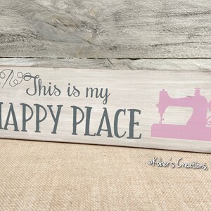 May include: A white wooden sign with the text "This is my HAPPY PLACE" in gray lettering. A pink silhouette of a sewing machine is on the right side of the sign. The sign is on a burlap background.