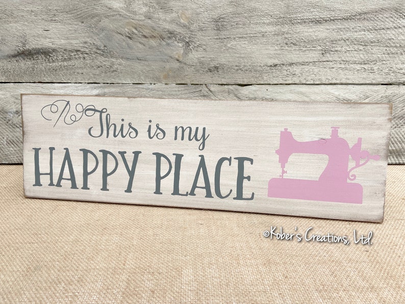 May include: A white wooden sign with the text "This is my HAPPY PLACE" in gray lettering. A pink silhouette of a sewing machine is on the right side of the sign. The sign is on a burlap background.