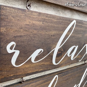 May include: A wooden sign with the word "relax" written in white cursive lettering. The sign is brown and has a distressed look. It is hanging from a hook.