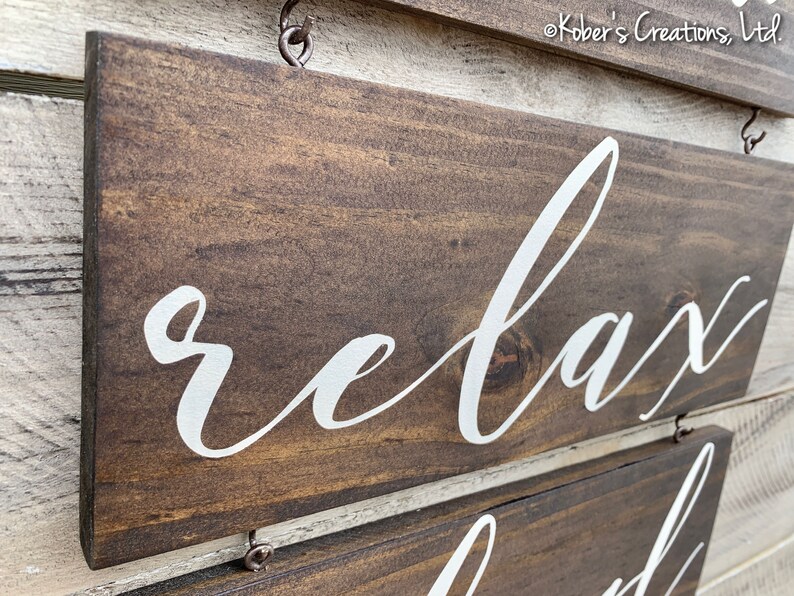 May include: A wooden sign with the word "relax" written in white cursive lettering. The sign is brown and has a distressed look. It is hanging from a hook.