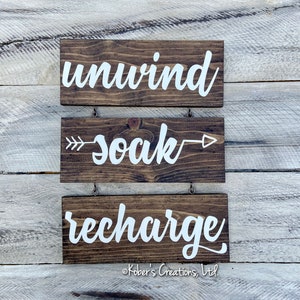 May include: Three wooden signs with white lettering hanging on a white wood wall. The top sign says "unwind", the middle sign says "soak" with arrows on either side, and the bottom sign says "recharge".