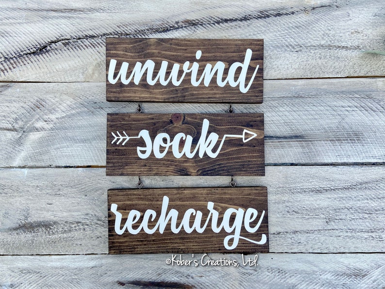 May include: Three wooden signs with white lettering hanging on a white wood wall. The top sign says "unwind", the middle sign says "soak" with arrows on either side, and the bottom sign says "recharge".