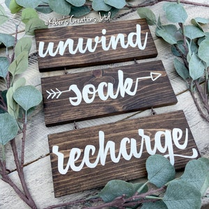 May include: Three wooden signs with white lettering hanging from twine. The top sign reads "unwind", the middle sign reads "soak" with an arrow pointing to the right, and the bottom sign reads "recharge".