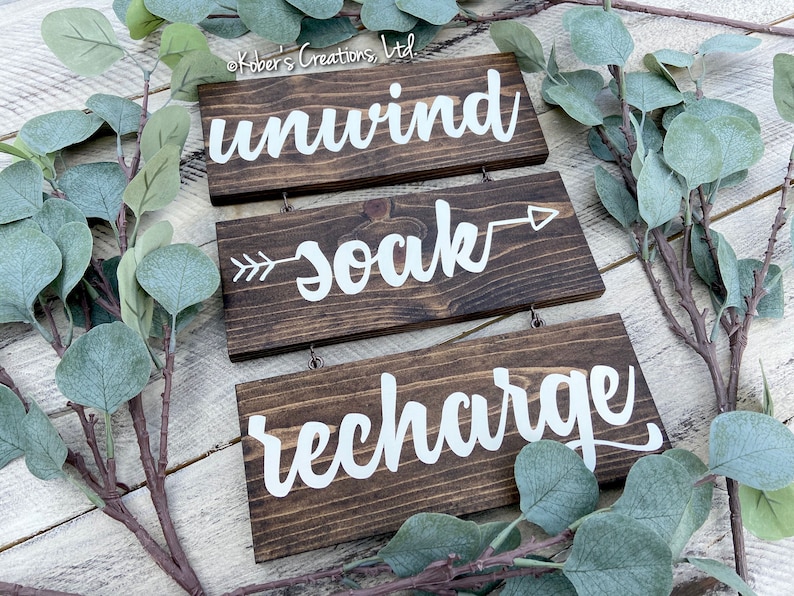 May include: Three wooden signs with white lettering hanging from twine. The top sign reads "unwind", the middle sign reads "soak" with an arrow pointing to the right, and the bottom sign reads "recharge".