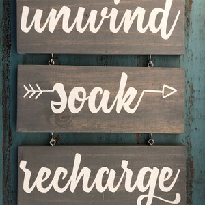 May include: Three wooden signs with white text. The top sign says "unwind", the middle sign says "soak" with arrows pointing left and right, and the bottom sign says "recharge".