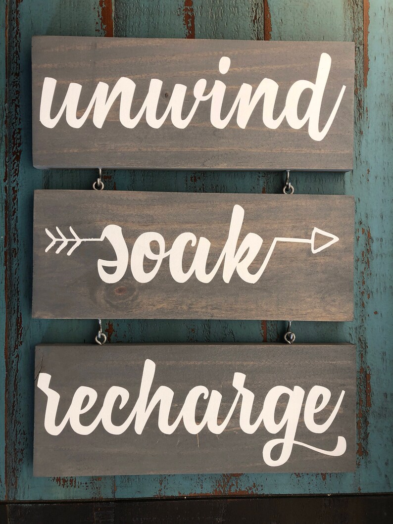May include: Three wooden signs with white text. The top sign says "unwind", the middle sign says "soak" with arrows pointing left and right, and the bottom sign says "recharge".