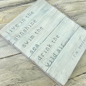 May include: A white wooden sign with a quote by R.W. Emerson: "Live in the sunshine. Swim the sea, drink the wild air."