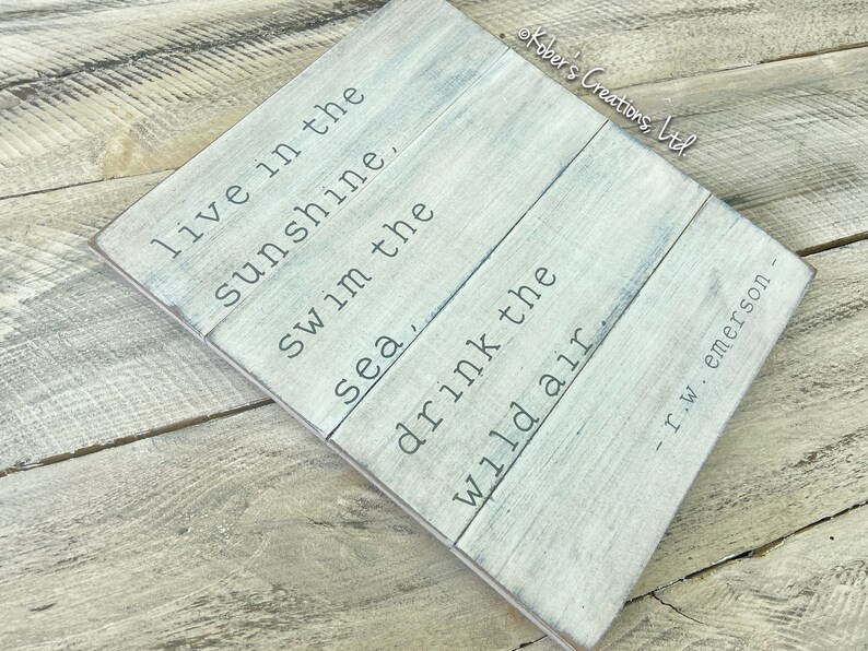 May include: A white wooden sign with a quote by R.W. Emerson: "Live in the sunshine. Swim the sea, drink the wild air."