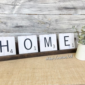 May include: White wooden tiles spelling out the word "HOME" with black lettering and a black number on each tile. The tiles are displayed on a dark brown wooden shelf. The shelf is sitting on a beige burlap surface.
