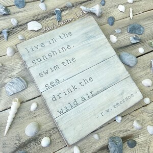 May include: A white wooden sign with a quote by R.W. Emerson: "live in the sunshine, swim the sea, drink the wild air." The sign is surrounded by seashells and stones on a wooden surface.