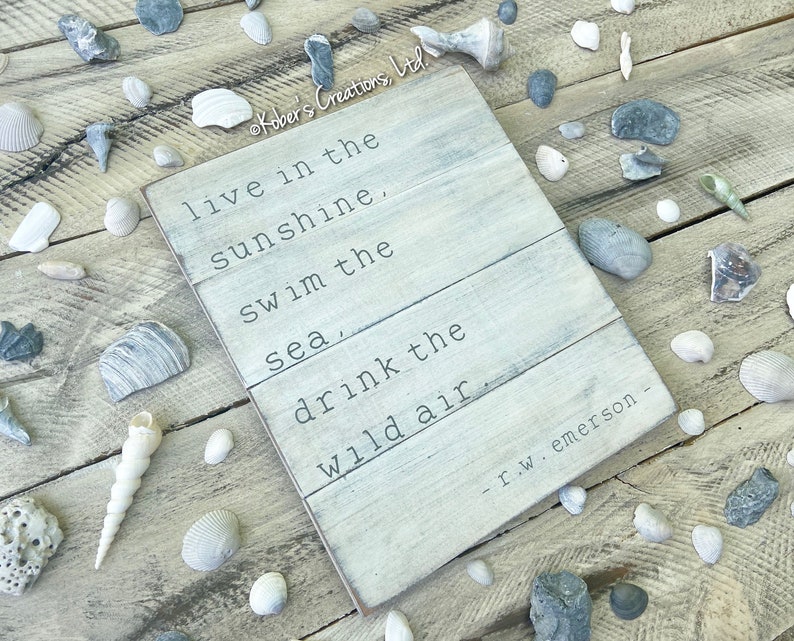 May include: A white wooden sign with a quote by R.W. Emerson: "live in the sunshine, swim the sea, drink the wild air." The sign is surrounded by seashells and stones on a wooden surface.
