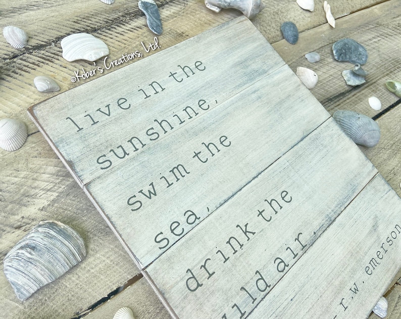 May include: A whitewashed wooden sign with a quote by Ralph Waldo Emerson: "Live in the sunshine, swim in the sea, drink the wild air."