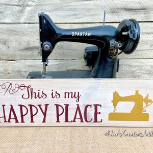 May include: A white wooden sign with the text "This is my HAPPY PLACE" in maroon lettering. A gold silhouette of a sewing machine is on the right side of the sign. The sign is sitting in front of a black vintage sewing machine with the word "SPARTAN" on the side.