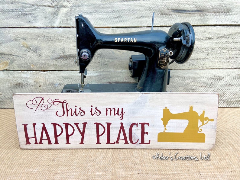 May include: A white wooden sign with the text "This is my HAPPY PLACE" in maroon lettering. A gold silhouette of a sewing machine is on the right side of the sign. The sign is sitting in front of a black vintage sewing machine with the word "SPARTAN" on the side.