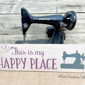 May include: A white wooden sign with the text "This is my HAPPY PLACE" in purple lettering. A stylized image of a sewing machine is on the right side of the sign. The sign is sitting in front of a vintage black sewing machine.