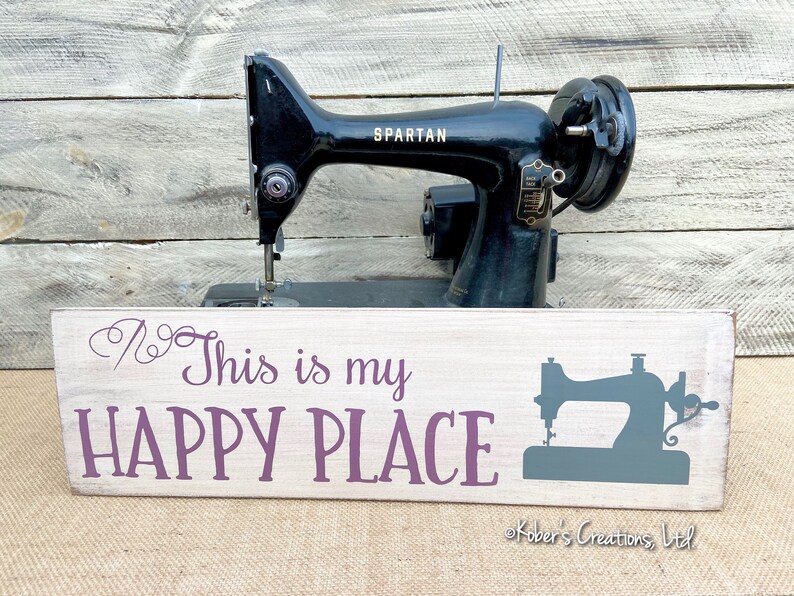 May include: A white wooden sign with the text "This is my HAPPY PLACE" in purple lettering. A stylized image of a sewing machine is on the right side of the sign. The sign is sitting in front of a vintage black sewing machine.