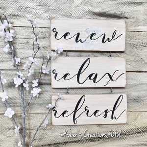 May include: Three white wooden signs with black calligraphy. The top sign says "renew", the middle sign says "relax", and the bottom sign says "refresh".