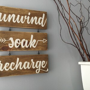May include: Three wooden signs with white painted text hang on a wall. The top sign says "unwind", the middle sign says "soak" with an arrow on each side, and the bottom sign says "recharge".