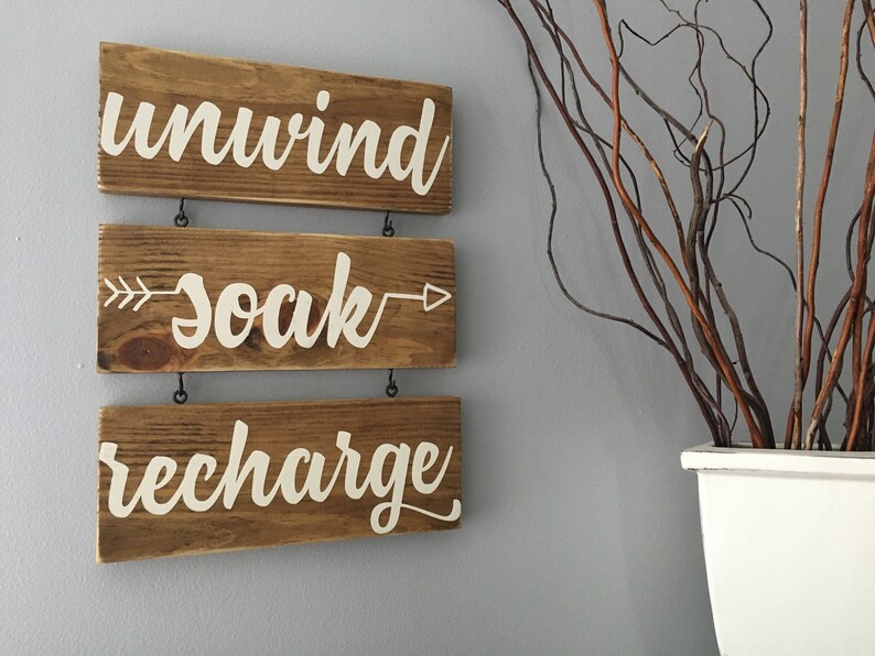 May include: Three wooden signs with white painted text hang on a wall. The top sign says "unwind", the middle sign says "soak" with an arrow on each side, and the bottom sign says "recharge".