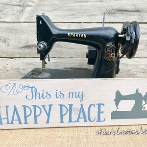 May include: White wooden sign with blue text that reads "This is my HAPPY PLACE" and a gray silhouette of a sewing machine. The sign is sitting in front of a vintage black sewing machine with the brand name "SPARTAN" on it. The sign is copyright "Kobers Creations, Ltd."