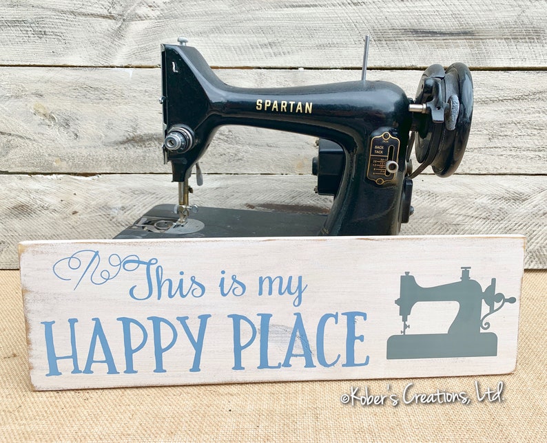 May include: White wooden sign with blue text that reads "This is my HAPPY PLACE" and a gray silhouette of a sewing machine. The sign is sitting in front of a vintage black sewing machine with the brand name "SPARTAN" on it. The sign is copyright "Kobers Creations, Ltd."
