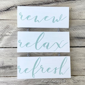 May include: Three white wooden signs with a light green cursive font. The top sign says "renew", the middle sign says "relax", and the bottom sign says "refresh".