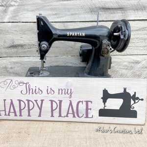 May include: A white wooden sign with purple text that reads "This is my HAPPY PLACE" and a black silhouette of a sewing machine. The sign is in front of a black vintage sewing machine with the word "SPARTAN" on it.