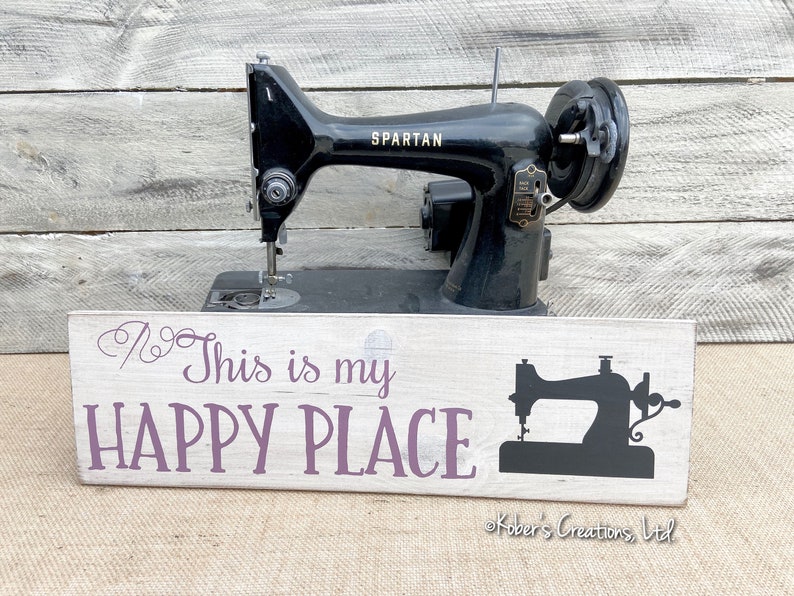 May include: A white wooden sign with purple text that reads "This is my HAPPY PLACE" and a black silhouette of a sewing machine. The sign is in front of a black vintage sewing machine with the word "SPARTAN" on it.