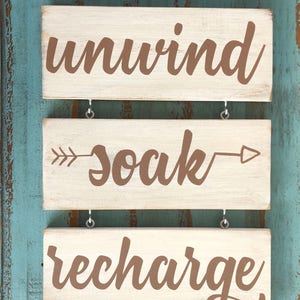 May include: Three wooden signs with white paint and brown lettering hanging on a blue wall. The top sign says "unwind", the middle sign says "soak" with arrows on either side, and the bottom sign says "recharge".