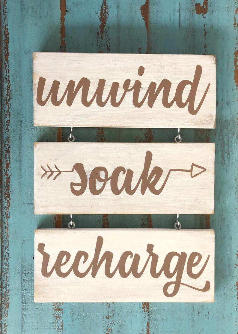 May include: Three wooden signs with white paint and brown lettering hanging on a blue wall. The top sign says "unwind", the middle sign says "soak" with arrows on either side, and the bottom sign says "recharge".