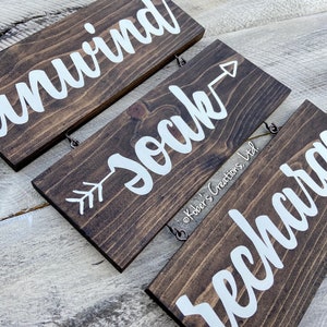 May include: Three wooden signs with white lettering hanging from a string. The top sign reads "unwind", the middle sign reads "soak" with an arrow pointing to the right, and the bottom sign reads "recharge".